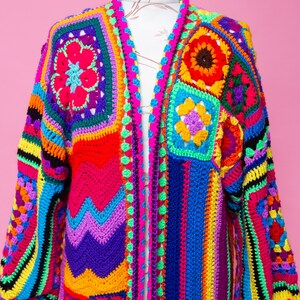 K&ouml;nnte beinhalten: Ein lebendiger, handgefertigter H&auml;kelcardigan in einem Patchwork-Design. Das farbenfrohe Kleidungsst&uuml;ck weist Quadrate und Streifen in Rot-, Blau-, Gelb-, Gr&uuml;n- und Lilat&ouml;nen auf. Der lang&auml;rmlige Cardigan wird auf einer Schaufensterpuppe vor rosa Hintergrund pr&auml;sentiert.