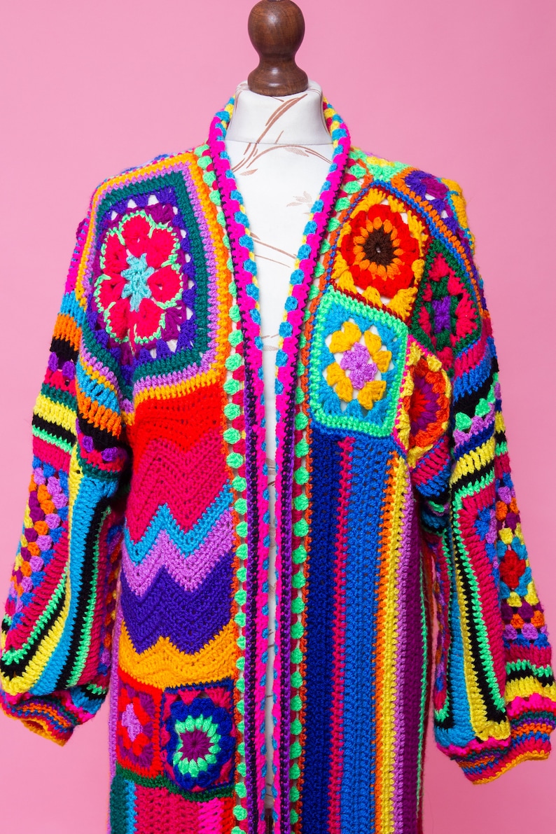 K&ouml;nnte beinhalten: Ein lebendiger, handgefertigter H&auml;kelcardigan in einem Patchwork-Design. Das farbenfrohe Kleidungsst&uuml;ck weist Quadrate und Streifen in Rot-, Blau-, Gelb-, Gr&uuml;n- und Lilat&ouml;nen auf. Der lang&auml;rmlige Cardigan wird auf einer Schaufensterpuppe vor rosa Hintergrund pr&auml;sentiert.