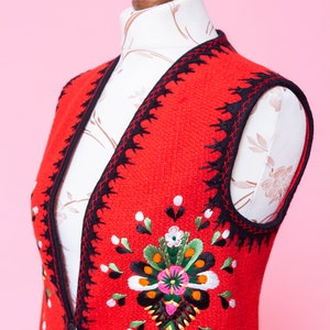 Pode incluir: Um colete vermelho sem mangas com acabamento preto e flores bordadas coloridas. O colete tem fecho de bot&atilde;o e bolsos.