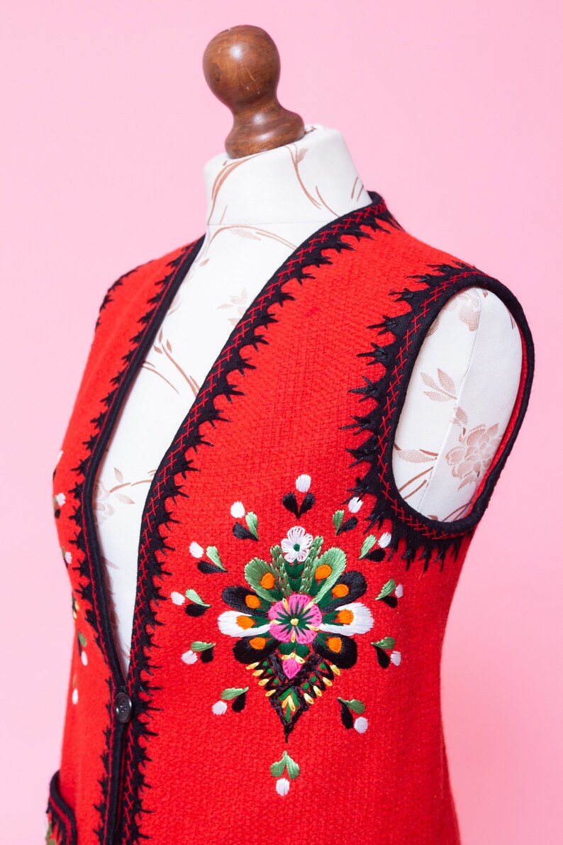 Pode incluir: Um colete vermelho sem mangas com acabamento preto e flores bordadas coloridas. O colete tem fecho de bot&atilde;o e bolsos.
