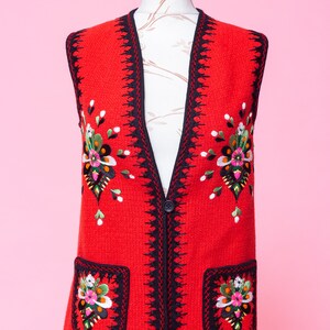 Pode incluir: Um colete vermelho sem mangas com guarni&ccedil;&atilde;o preta e bordados florais coloridos. O colete tem dois bolsos com flores bordadas na frente.