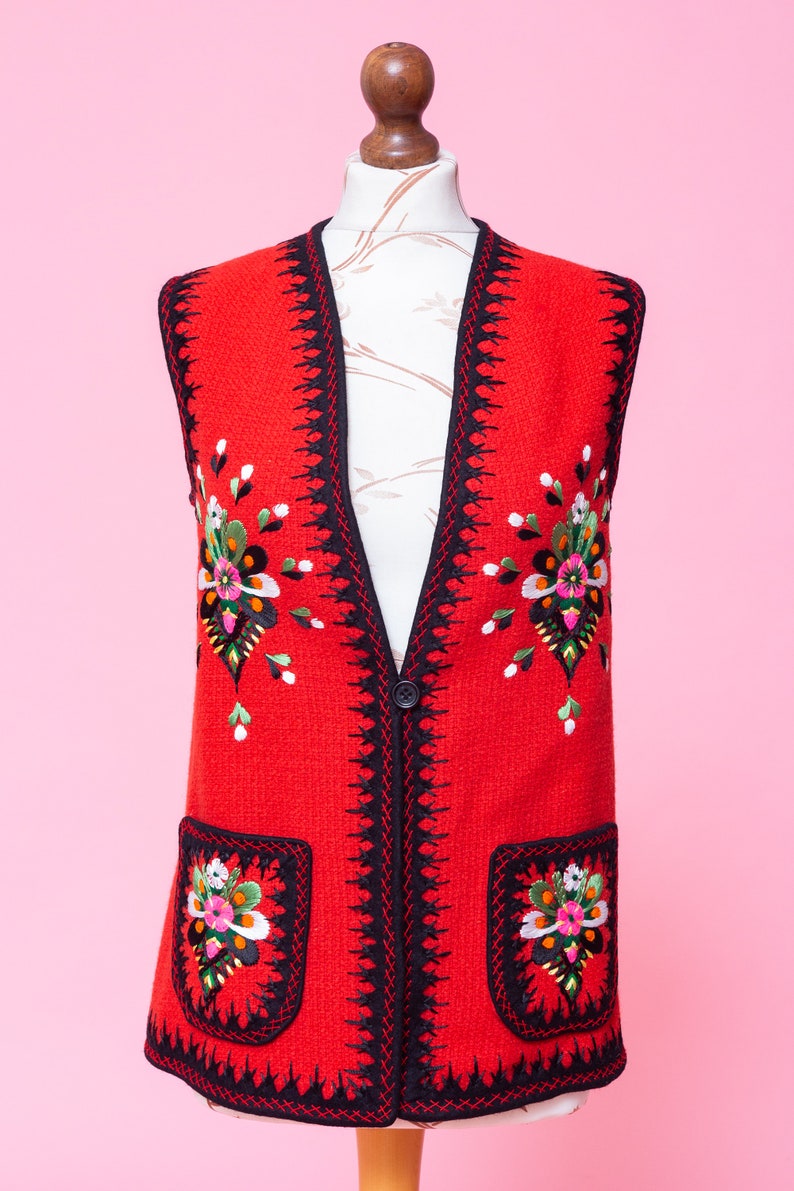 Pode incluir: Um colete vermelho sem mangas com guarni&ccedil;&atilde;o preta e bordados florais coloridos. O colete tem dois bolsos com flores bordadas na frente.