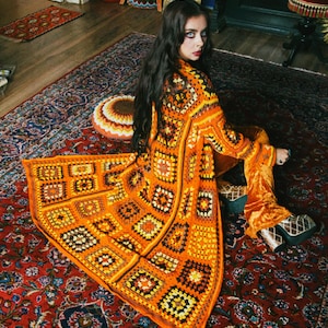 Puede incluir: Una mujer lleva una larga chaqueta de ganchillo naranja, marrón y amarilla con un patrón geométrico. Está sentada en una alfombra roja con estampado.