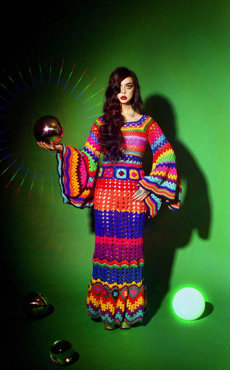 May include: A vibrant, handmade crochet dress in a rainbow of colors, featuring long bell sleeves and a floor-length skirt. The dress is worn by a person holding a gold sphere. The background is a solid green.