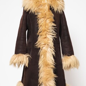 May include: A dark brown coat with a tan faux fur trim and lining. The coat features a hood, cuffs, and a front trim of long, shaggy faux fur. The coat is displayed on a wooden mannequin.