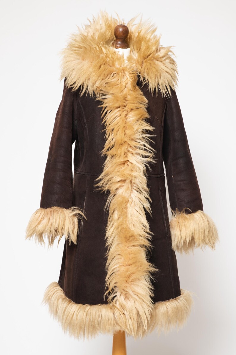 May include: A dark brown coat with a tan faux fur trim and lining. The coat features a hood, cuffs, and a front trim of long, shaggy faux fur. The coat is displayed on a wooden mannequin.