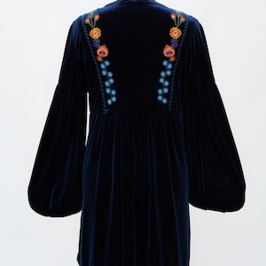 May include: A navy blue velvet dress with long, balloon sleeves. The dress has colourful floral embroidery on the back and shoulders. It is displayed on a mannequin against a white background.