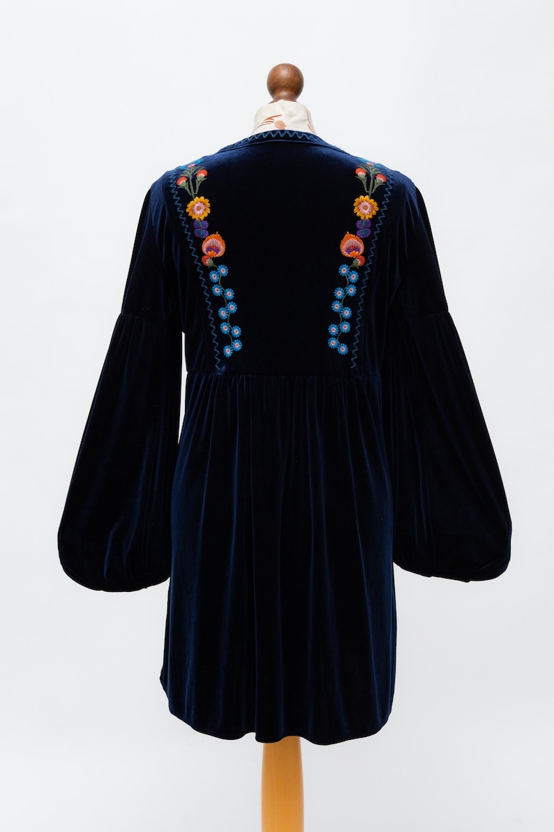 May include: A navy blue velvet dress with long, balloon sleeves. The dress has colourful floral embroidery on the back and shoulders. It is displayed on a mannequin against a white background.