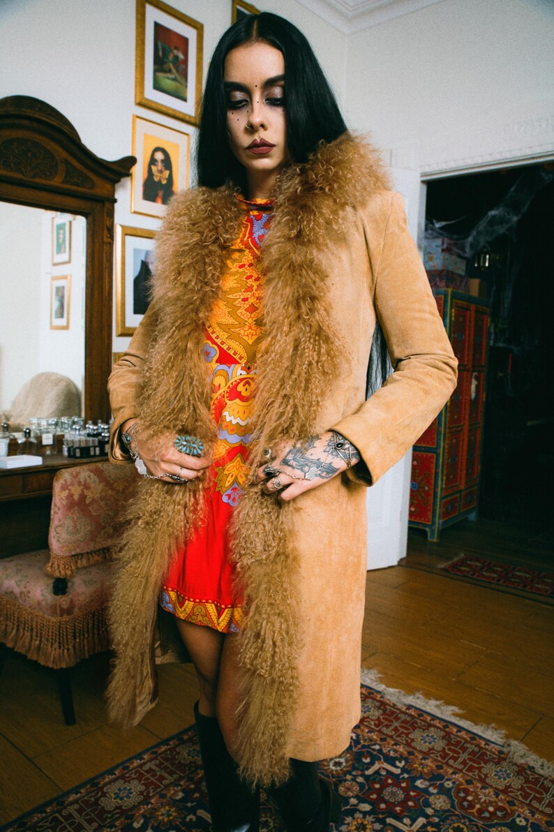 May include: A tan suede coat with a shaggy fur collar and trim. The coat is worn over a red and orange patterned dress. The image is taken indoors, with a vintage aesthetic. The coat is knee-length and has a classic silhouette.