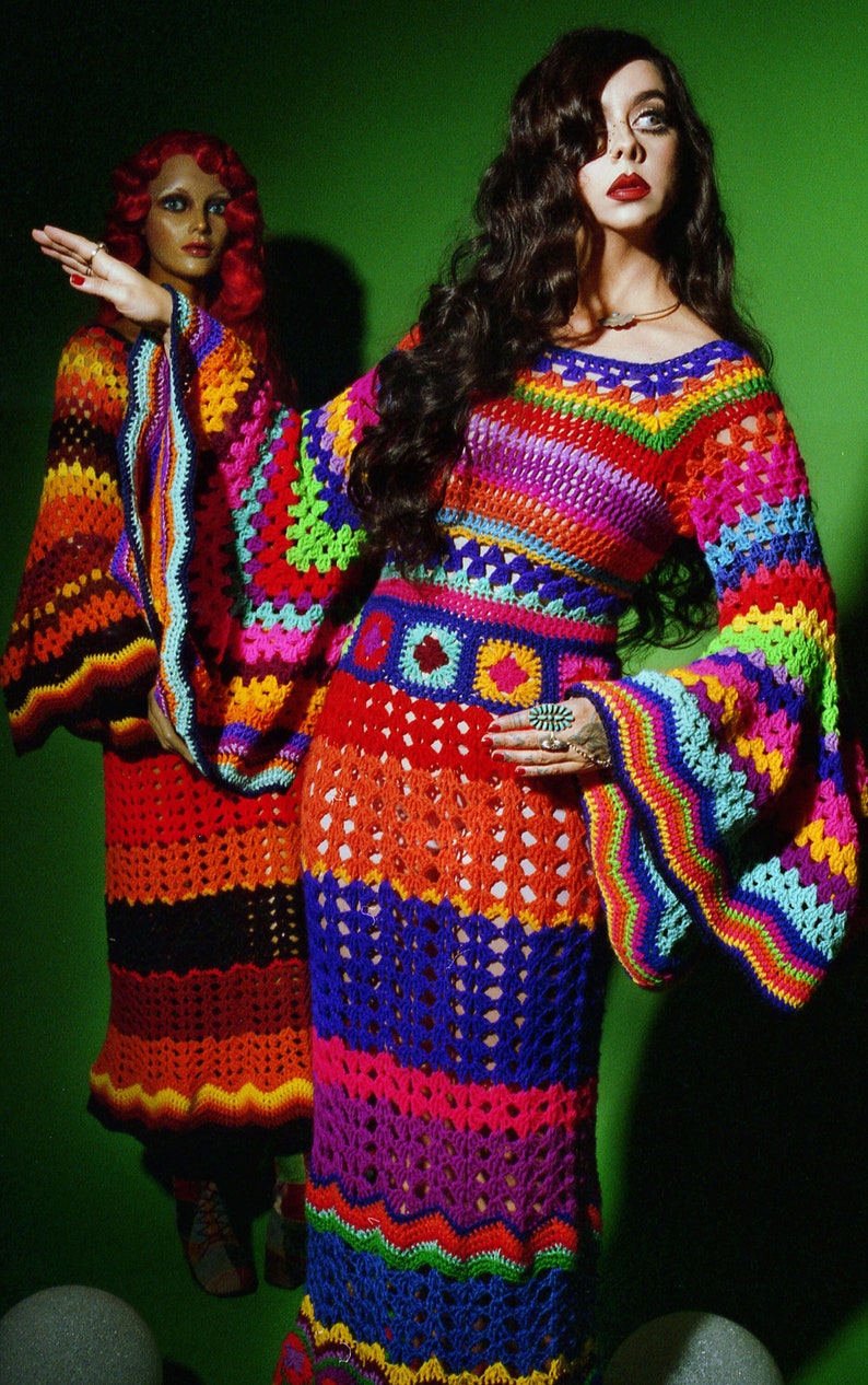 May include: A vibrant, handmade crochet dress in a rainbow of colors, featuring long bell sleeves and a fitted bodice. The dress has a unique openwork design and is worn by a person with long, dark hair. A second dress is visible in the background.