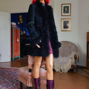 May include: A mannequin wearing a black suede coat with feather trim, a short pink dress, and tall purple lace-up boots. The mannequin has red curly hair and is standing in a room with a patterned rug and antique furniture.