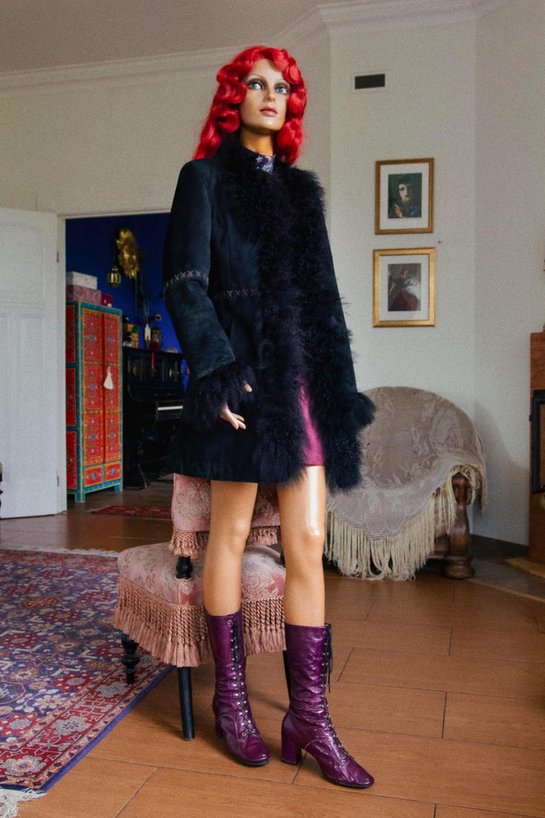 May include: A mannequin wearing a black suede coat with feather trim, a short pink dress, and tall purple lace-up boots. The mannequin has red curly hair and is standing in a room with a patterned rug and antique furniture.