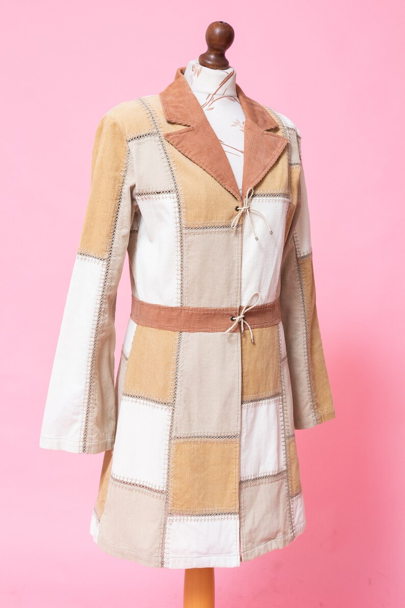 K&ouml;nnte beinhalten: Eine lange Patchworkjacke mit einem braunen Cordkragen und einem braunen G&uuml;rtel. Die Jacke ist aus verschiedenen Beige- und Wei&szlig;t&ouml;nen aus Cord gefertigt.