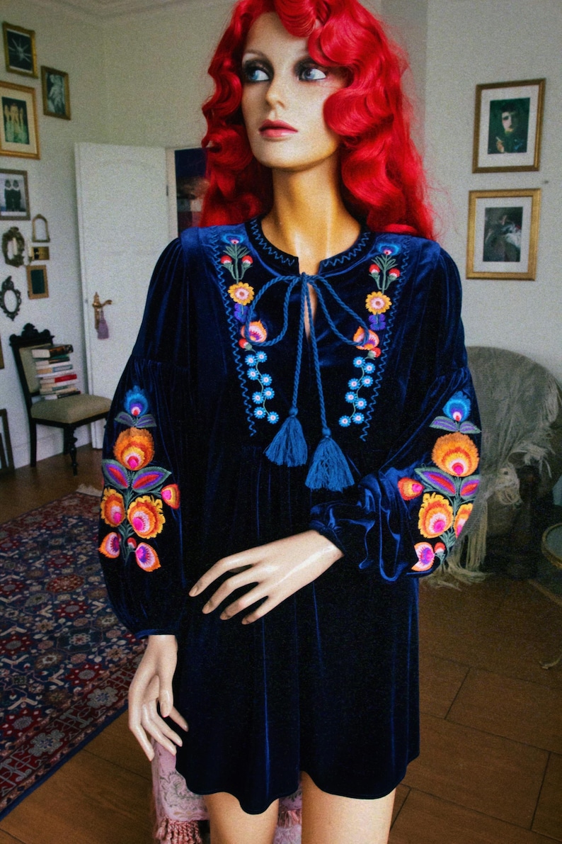 May include: A dark blue velvet dress with long sleeves and colourful floral embroidery on the neckline and sleeves. The dress has a tie closure at the neckline and is displayed on a mannequin with red hair.