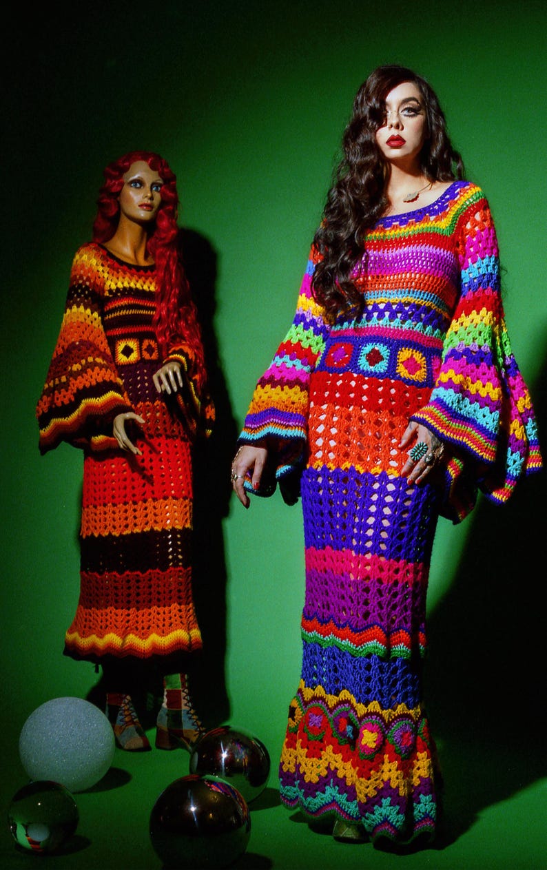 May include: Two women's crochet dresses in vibrant rainbow colors. One dress is on a mannequin, the other is worn by a person. The dresses have bell sleeves and a long, flowing skirt. The background is a solid green.