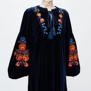 May include: A navy blue velvet dress with vibrant floral embroidery on the sleeves and neckline. The dress has long, balloon sleeves and a tie-neck with tassels. The embroidery features orange, pink, blue, and yellow flowers.