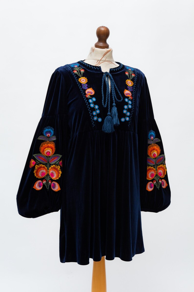 May include: A navy blue velvet dress with vibrant floral embroidery on the sleeves and neckline. The dress has long, balloon sleeves and a tie-neck with tassels. The embroidery features orange, pink, blue, and yellow flowers.