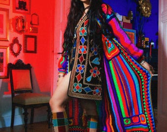 Pode incluir: Um traje boêmio colorido com um casaco longo de crochê aberto com um design de listras de arco-íris. O traje inclui um vestido preto bordado e botas de patchwork. O quarto tem paredes vermelhas e uma estética vintage.