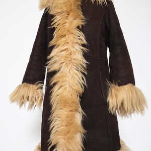 May include: A dark brown coat with a tan faux fur collar, trim, and cuffs. The coat is displayed on a wooden mannequin. The coat has long sleeves and a knee-length hem. The faux fur trim adds a luxurious touch to this outerwear.