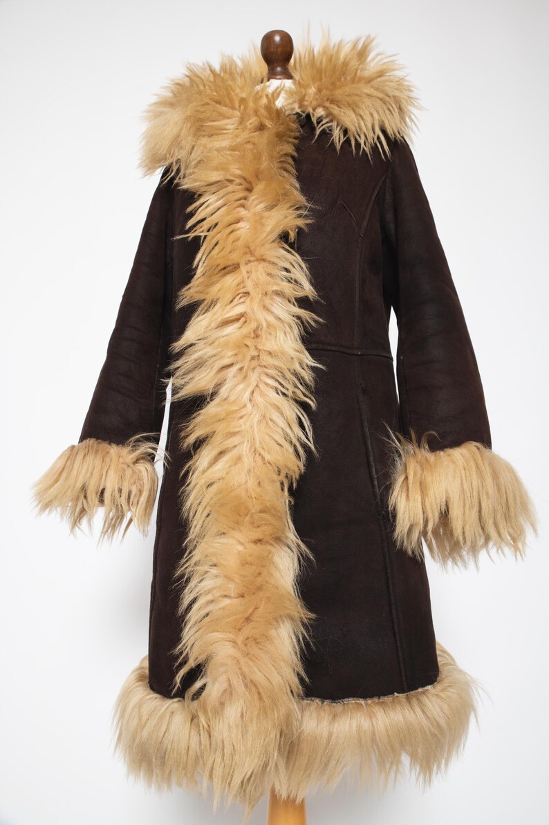 May include: A dark brown coat with a tan faux fur collar, trim, and cuffs. The coat is displayed on a wooden mannequin. The coat has long sleeves and a knee-length hem. The faux fur trim adds a luxurious touch to this outerwear.