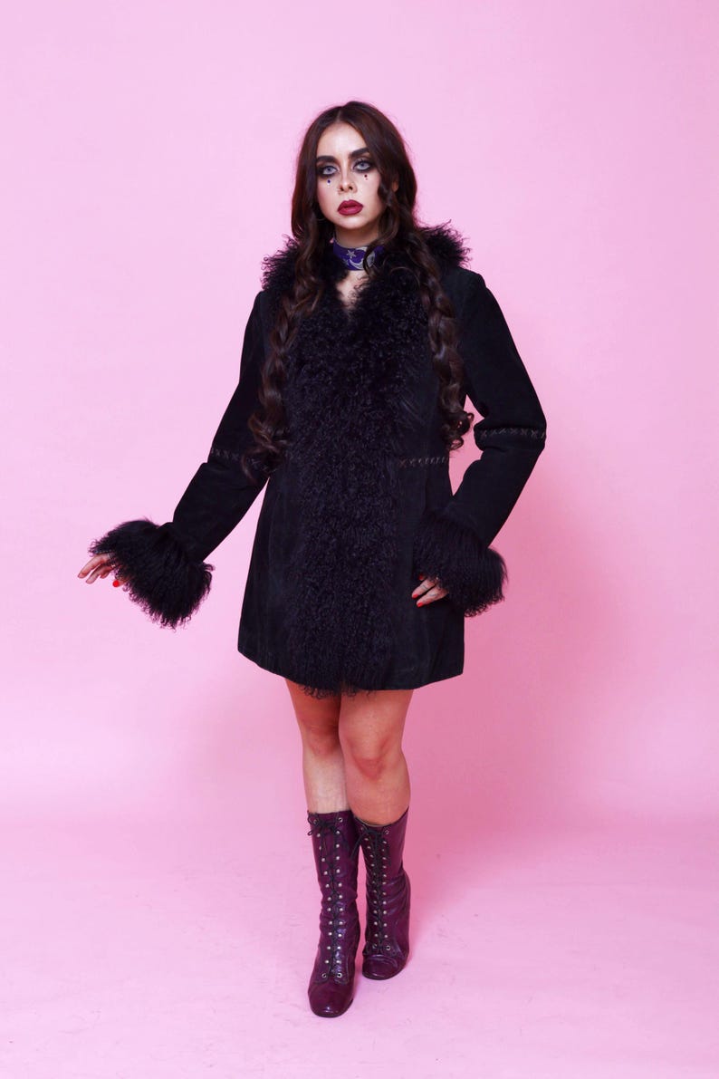 May include: Black suede coat with faux fur trim on the collar, cuffs, and front. The coat has a flared silhouette and long sleeves. The model is wearing a purple choker and purple lace-up boots. The background is a solid pink.