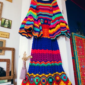 May include: A vibrant, handmade crochet dress in a rainbow of colors, including blue, red, yellow, and green. The dress features long sleeves with flared cuffs and a full skirt. It is hanging on a wooden hanger against a white door.