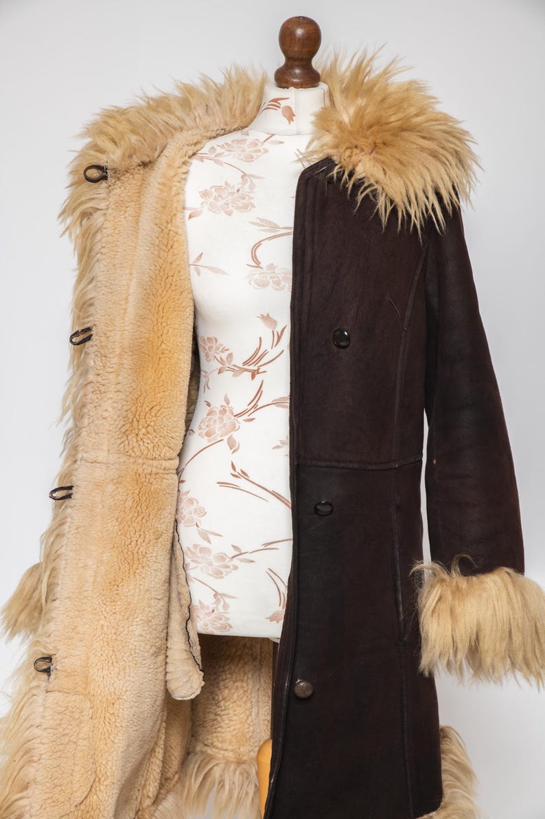 May include: A vintage brown shearling coat with a tan fur collar and cuffs. The coat is buttoned and displayed on a mannequin. The interior lining is a light tan color, and the exterior is a dark brown. The coat has a classic, retro style.