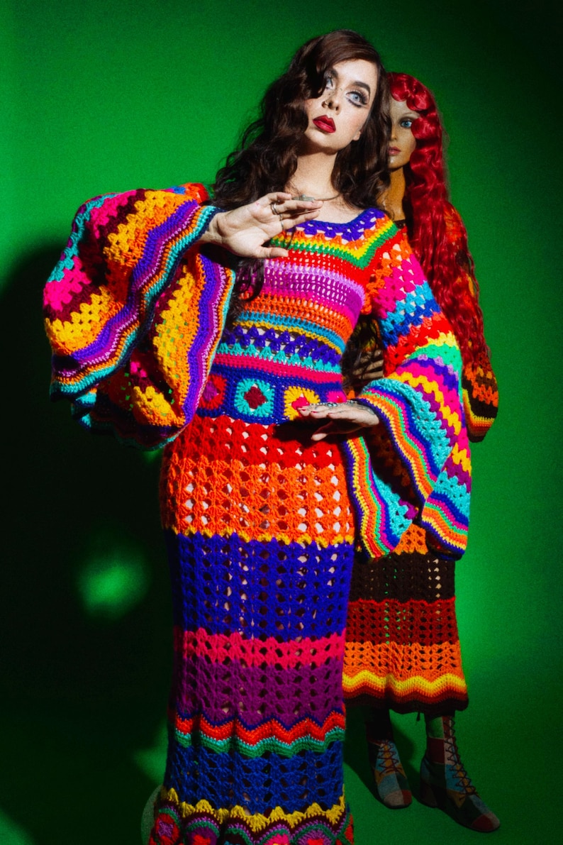 May include: A vibrant, handmade crochet dress in a rainbow of colors, including pink, orange, yellow, blue, and purple. The dress features long, flared sleeves and a fitted bodice, with a skirt that flares out. The dress is a unique, eye-catching piece.