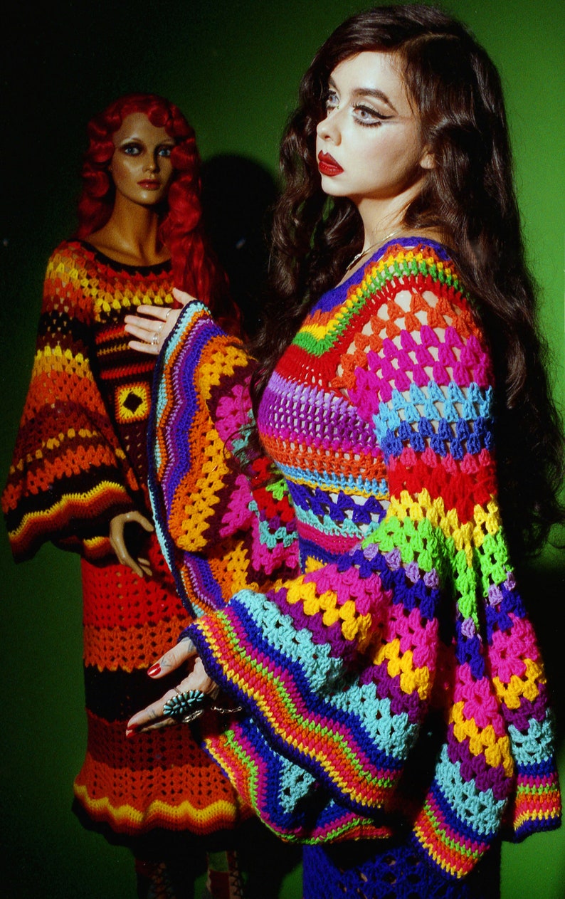 May include: A vibrant, rainbow-colored crochet top with flared sleeves. The top features a mix of bright colors including pink, blue, yellow, and green. A mannequin in the background wears a similar crochet dress in shades of orange and red.