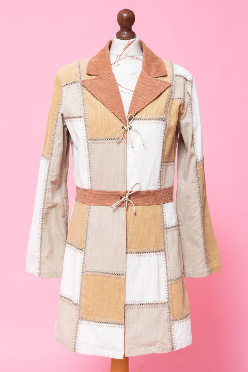 K&ouml;nnte beinhalten: Eine lang&auml;rmelige Patchworkjacke mit braunem Cordkragen und G&uuml;rtel. Die Jacke ist aus beigefarbenem, braunem und wei&szlig;em Stoff.