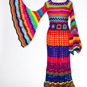 May include: A vibrant, handmade crochet dress in a rainbow of colors. The long dress features bell sleeves and a flared skirt. The bodice and sleeves are striped with various colors, while the skirt has a mix of solid and openwork sections. The dress is displayed on a mannequin.