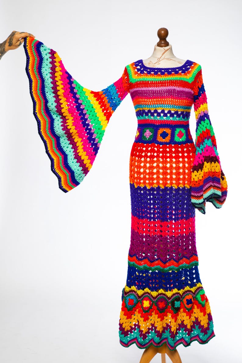 May include: A vibrant, handmade crochet dress in a rainbow of colors. The long dress features bell sleeves and a flared skirt. The bodice and sleeves are striped with various colors, while the skirt has a mix of solid and openwork sections. The dress is displayed on a mannequin.