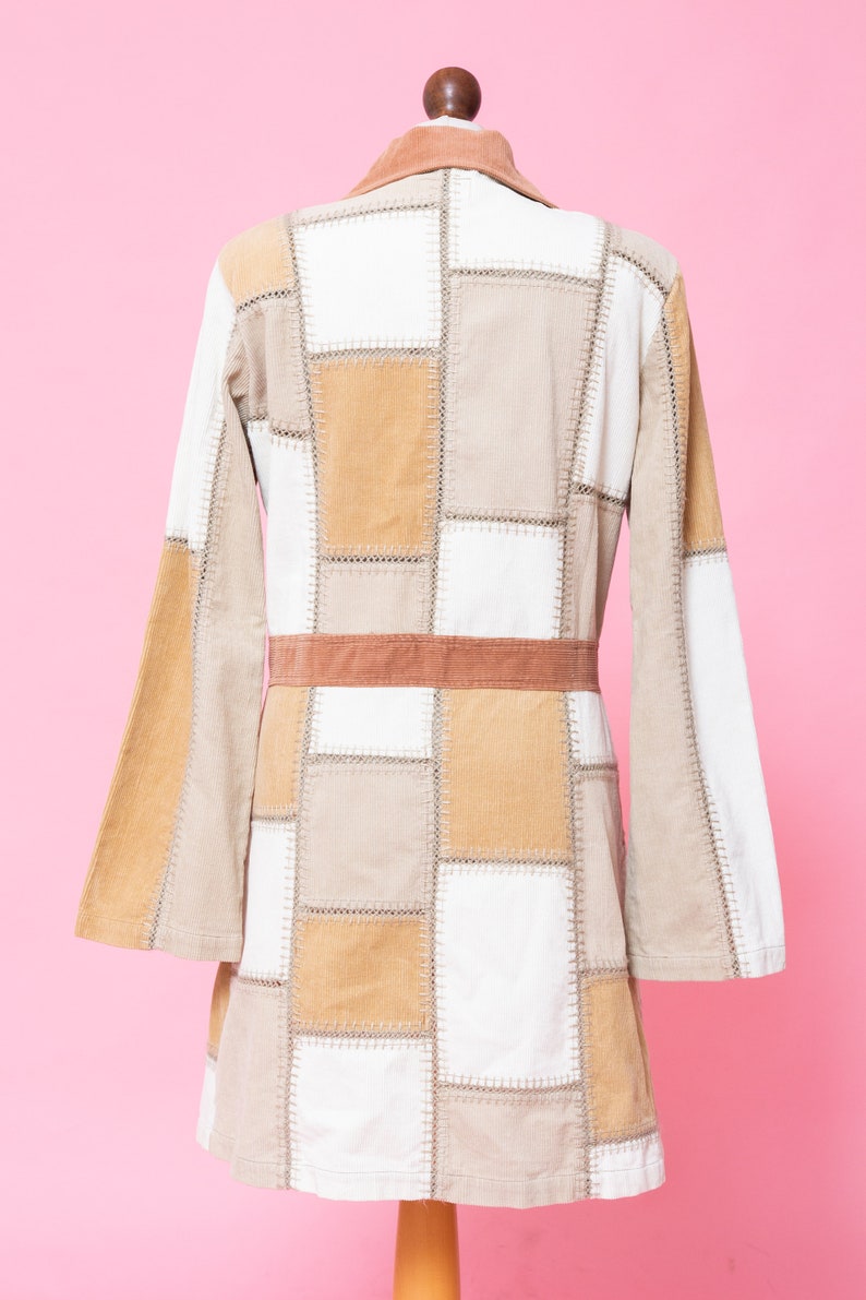 K&ouml;nnte beinhalten: Eine Patchworkjacke mit braunem Cordkragen und G&uuml;rtel. Die Jacke ist aus beigefarbenem und wei&szlig;em Cord und hat ein quadratisches Patchworkmuster.