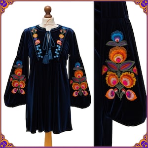 May include: A navy blue velvet dress with long, bell sleeves. The dress features colourful floral embroidery in shades of orange, pink, blue, and green on the sleeves and bodice. It has a round neckline with a tassel tie.