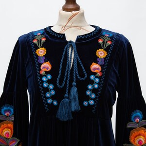 May include: A navy blue velvet dress with vibrant floral embroidery. The dress has long sleeves with bell cuffs and a tie-neck with tassels. The embroidery features orange, yellow, blue, and purple flowers.