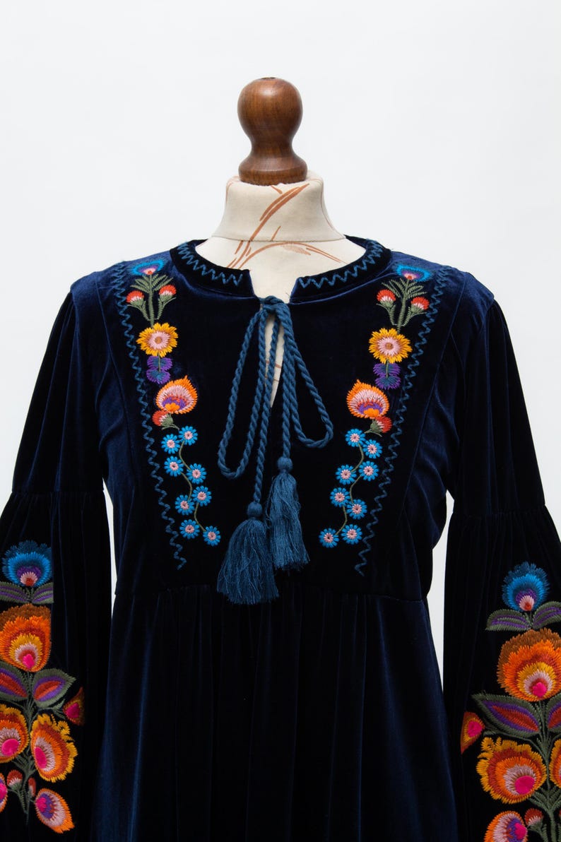 May include: A navy blue velvet dress with vibrant floral embroidery. The dress has long sleeves with bell cuffs and a tie-neck with tassels. The embroidery features orange, yellow, blue, and purple flowers.