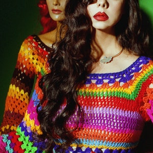 May include: A colorful crochet top and skirt set in a rainbow of hues, including red, orange, yellow, green, blue, and purple. The top has long sleeves and a square neckline. The model is wearing a silver leaf pendant necklace and rings.
