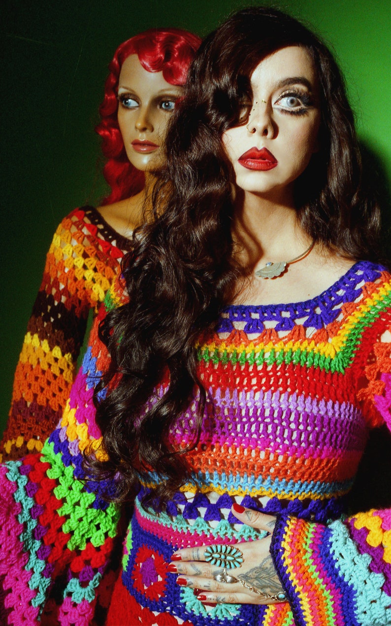 May include: A colorful crochet top and skirt set in a rainbow of hues, including red, orange, yellow, green, blue, and purple. The top has long sleeves and a square neckline. The model is wearing a silver leaf pendant necklace and rings.