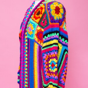 K&ouml;nnte beinhalten: Ein lebendiger, handgefertigter H&auml;kelcardigan im Patchwork-Design. Der lange, offen geschnittene Pullover hat einen Farbenmix aus Pink, Blau, Gelb, Gr&uuml;n und Lila. Das Kleidungsst&uuml;ck wird auf einer Schaufensterpuppe vor einem rosa Hintergrund pr&auml;sentiert.