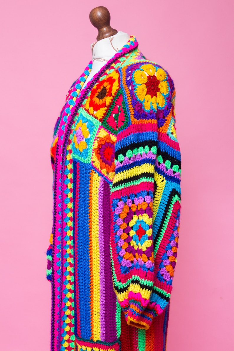 K&ouml;nnte beinhalten: Ein lebendiger, handgefertigter H&auml;kelcardigan im Patchwork-Design. Der lange, offen geschnittene Pullover hat einen Farbenmix aus Pink, Blau, Gelb, Gr&uuml;n und Lila. Das Kleidungsst&uuml;ck wird auf einer Schaufensterpuppe vor einem rosa Hintergrund pr&auml;sentiert.