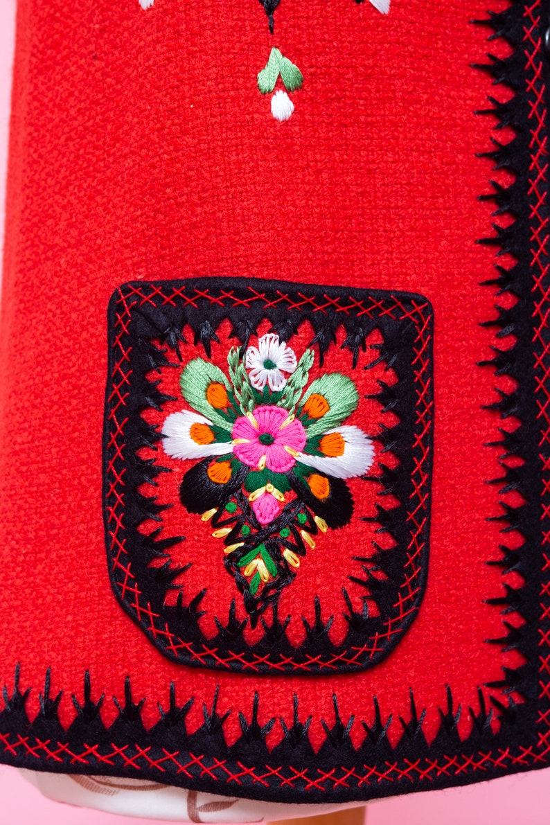 Pode incluir: Tecido vermelho com acabamento bordado preto e um desenho floral no centro do tecido. O desenho floral &eacute; bordado em tons de rosa, verde, branco e laranja.