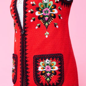 Pode incluir: Um colete vermelho sem mangas com guarni&ccedil;&atilde;o preta e desenhos florais bordados. O colete tem dois bolsos com desenhos florais bordados.