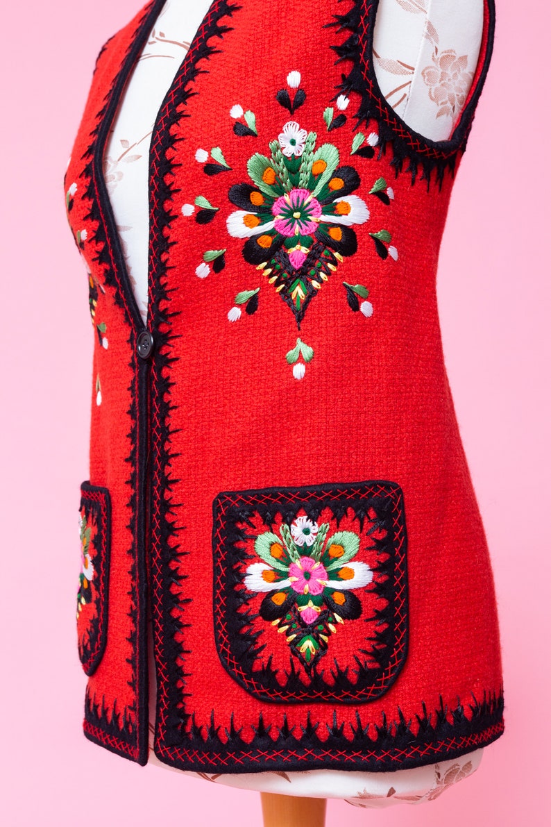Pode incluir: Um colete vermelho sem mangas com guarni&ccedil;&atilde;o preta e desenhos florais bordados. O colete tem dois bolsos com desenhos florais bordados.