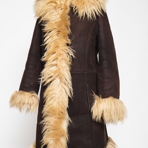 May include: A dark brown coat with a tan faux fur collar, cuffs, and trim. The coat is displayed on a wooden mannequin. The coat has a long length and a classic design, suitable for cold weather.