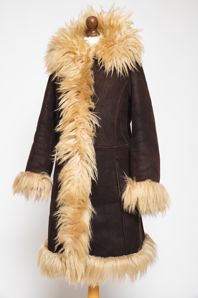 May include: A dark brown coat with a tan faux fur collar, cuffs, and trim. The coat is displayed on a wooden mannequin. The coat has a long length and a classic design, suitable for cold weather.
