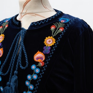 May include: A dark blue velvet top with colourful floral embroidery. The embroidery features orange, yellow, blue, and purple flowers. The top has a round neckline with a blue cord and tassels.