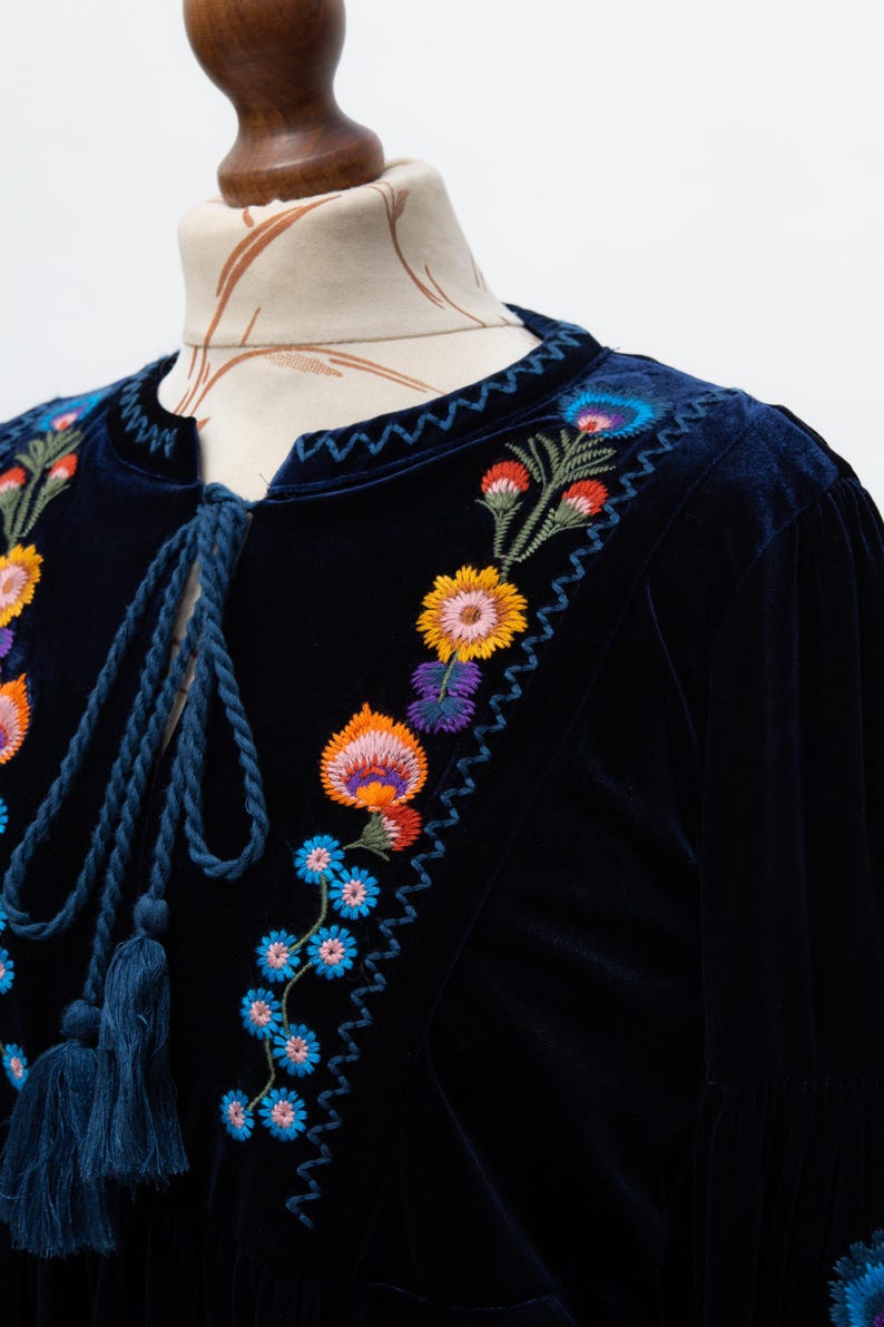 May include: A dark blue velvet top with colourful floral embroidery. The embroidery features orange, yellow, blue, and purple flowers. The top has a round neckline with a blue cord and tassels.