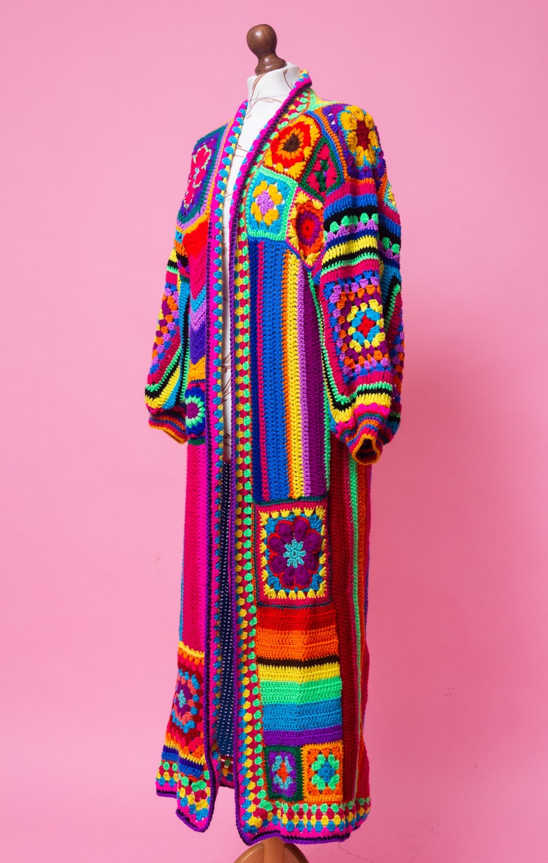 K&ouml;nnte beinhalten: Lange, bunte H&auml;keljacke auf einer Schaufensterpuppe. Das Kleidungsst&uuml;ck hat ein Patchwork-Design mit Quadraten und Streifen in Rot, Orange, Gelb, Gr&uuml;n, Blau und Lila. Es hat lange &Auml;rmel und einen Kragen.