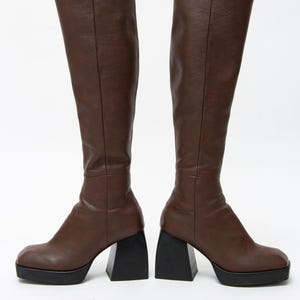 Puede incluir: Un par de botas marrones hasta la rodilla con suela de plataforma y tacón negro grueso. Las botas tienen una textura lisa similar al cuero y una punta redondeada. El tacón mide aproximadamente 10 cm.