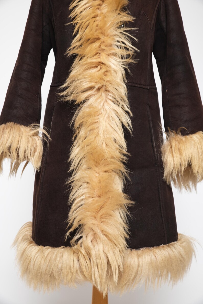 May include: A dark brown coat with long sleeves and a faux fur trim. The coat features a light brown faux fur collar, cuffs, and hem. The coat is displayed on a wooden stand against a white background.